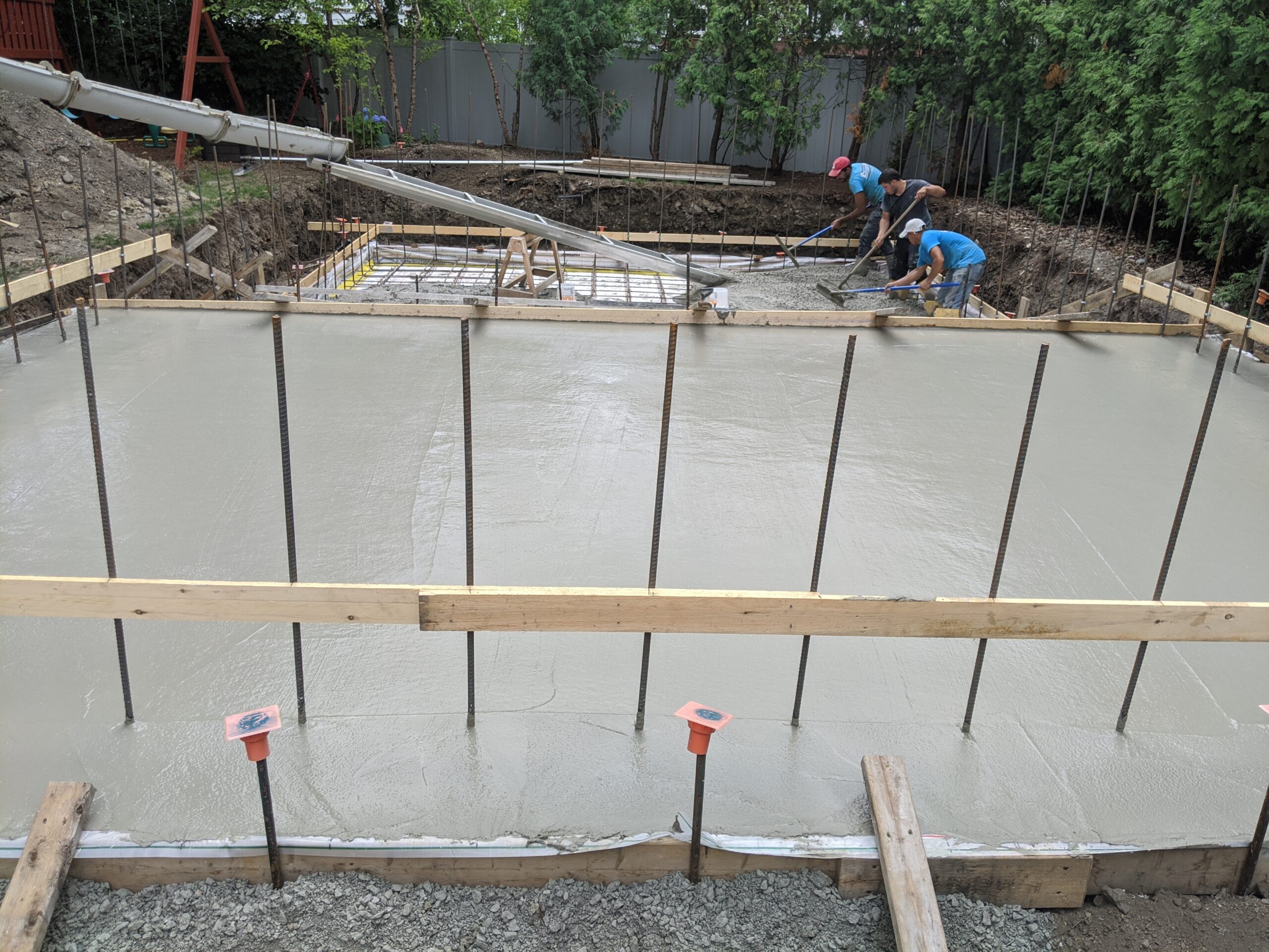 a vew from the top of the pool as concrete is poured into the base layer of the pool which will be built with ICF forms. a team of workers lays out the concrete at the lower end of the pool.