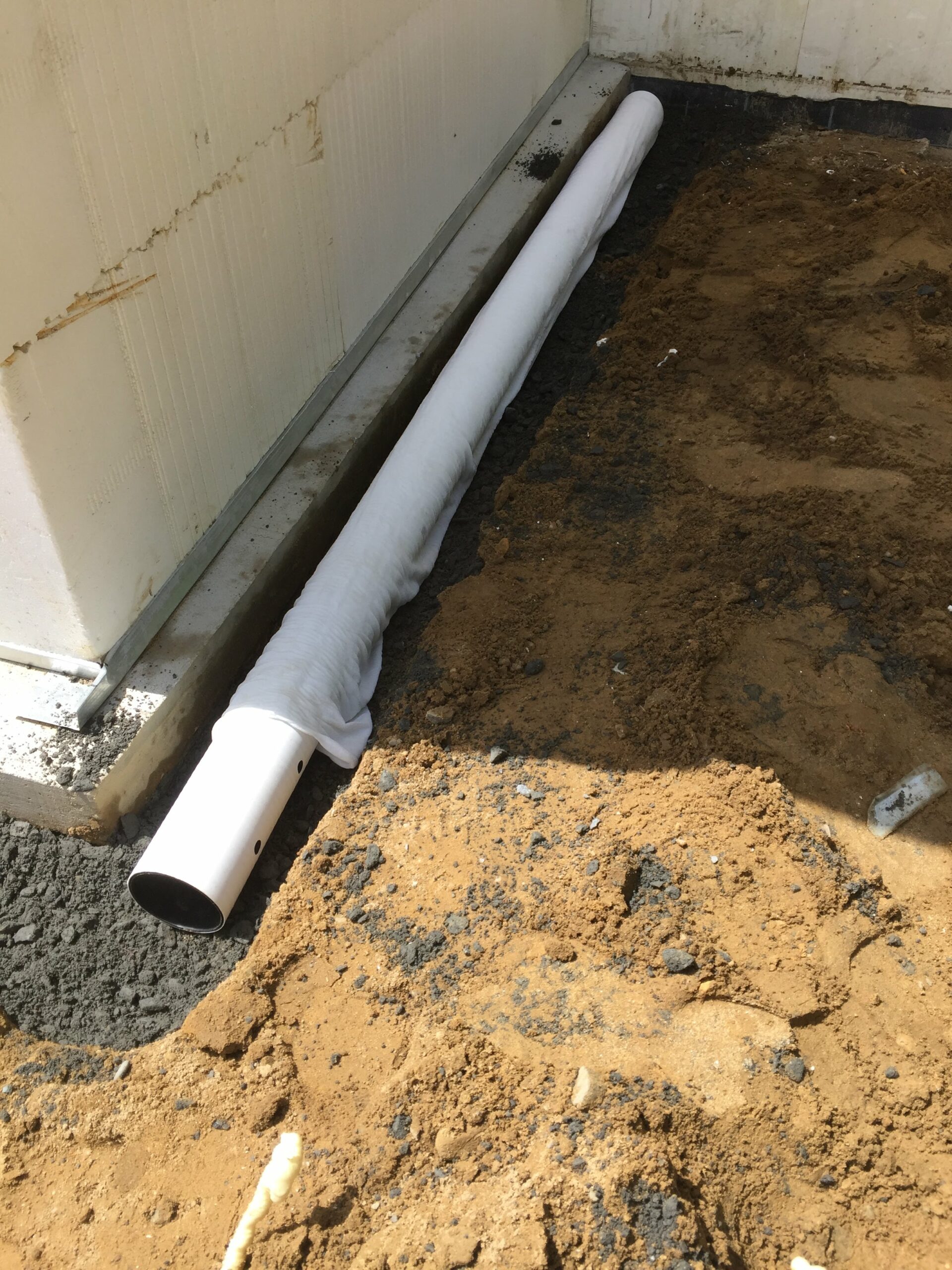 a pipe is seen next to the foundation of a building. the pipe has holes drilled into it and a layer of permeable material over the top covers the pipe which will be covered by gravel to provide a waterproofing mechanism to the foundation of the ICF home.
